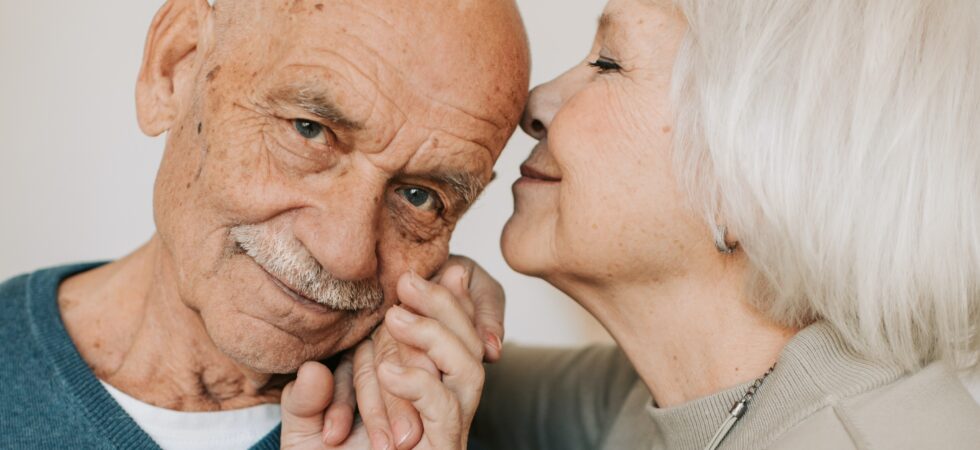 We see an older couple standing in front of a cream colored wall. The man is completely bald with a mustache and wears a teal blue V-necked pullover sweater over a white tee shirt. He leans his head toward an older woman with a short, white bob. We see her only in profile as she leans toward kissing the older man's temple. She wears a beighbe twinset. Both show gentle smiles.