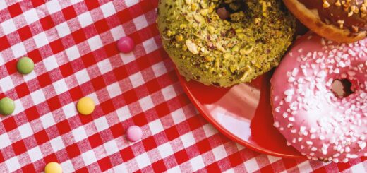 We see a red and white gingham table cloth spread out with a read plate of donuts. The visible donuts have sprinkles and frosting, one peanut butter, one chocolate, and one strawberry.
