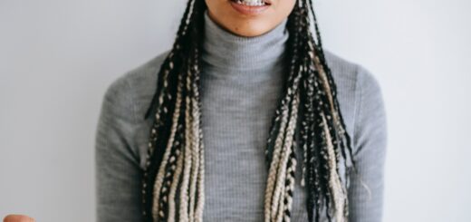 This indoor image shows a woman with long, intricately braided gray hair, some braided on top and some loose over her shoulders. She's wearing a gray turtleneck sweater. Her expression shows her explaining something to a person off camera and she holds her hands out, palm open upward.