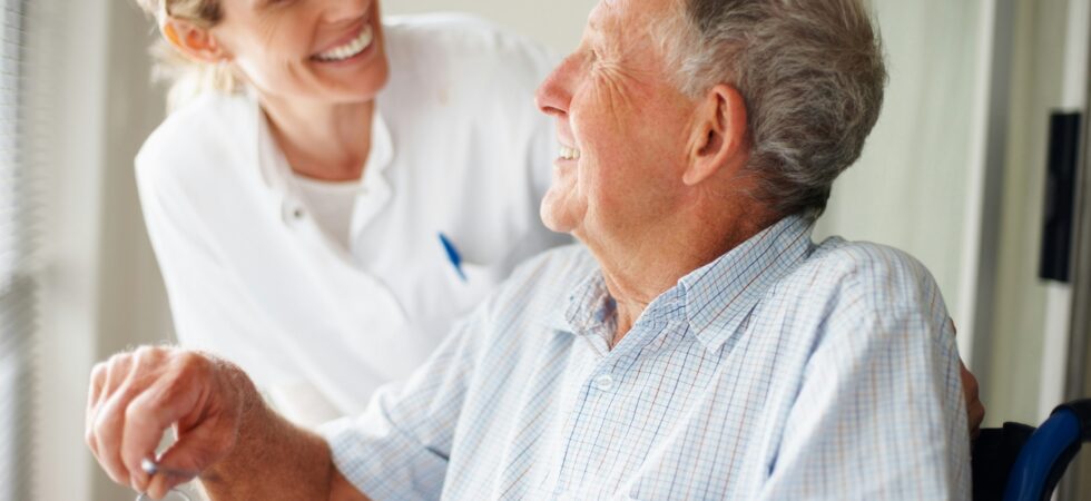 This indoor image shows an older man in the forground. He's sitting in a chair but looking up at a younger woman who is smiling at him. He wears a pastel plaid summer sport shirt and she wears a white professional jacket.