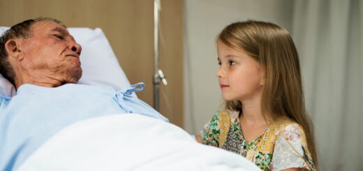 This image shows and older man lying on a hospital bed. A young child with should-length brown hair and a print top looks at him as he gazes back.