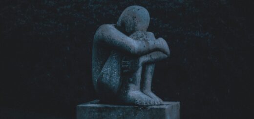 This nighttime image shows a stone statue sitting on top of a low pillar support. The statue is sitting with knees drawn in close and arms laying atop the knees. The head is bowed.