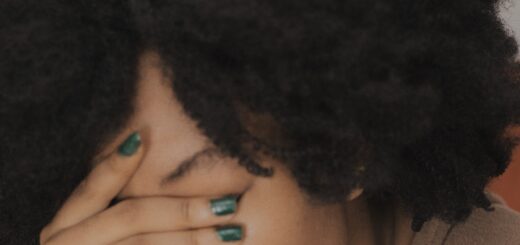 This very close image shows a woman who has her open hand over her eyes as she looks down as if leaning on something. She is wearing blue nailpolish and a beighe top