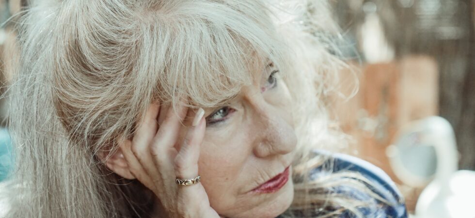 This outdoor image shows a woman with very blonde, poofy hair pulled back and we see a fall of hair behind her head. She's leaning her head sideways supporting it with her hand as she looks to the side. She is wearing a sliver pinky ring and pink nail polish, on her fingers and has on a blue plaid cotton type shirt.