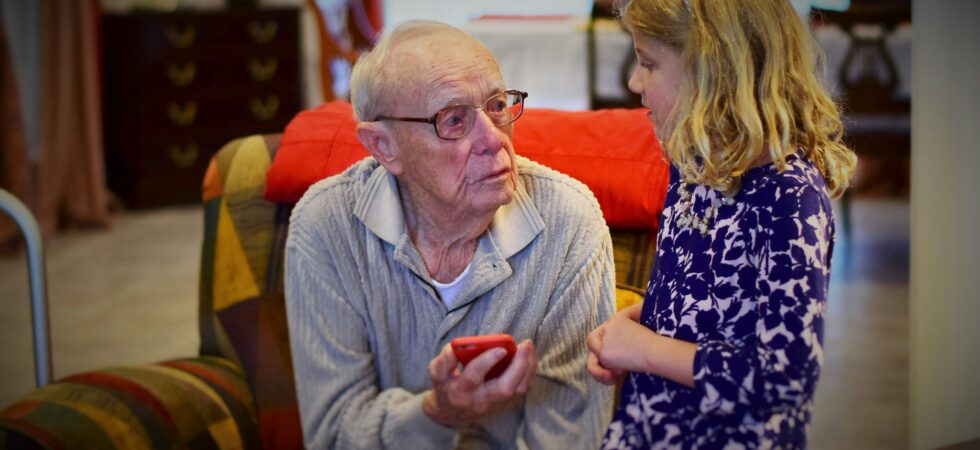 This indoor image shows an older man with receding white hair, wearing a gray shirt with an open collar. He holds a cell phone in one hand as he looks up at a young girl with long dark blonde hair. She wears a purple print top and appears to be explaining something to the man.