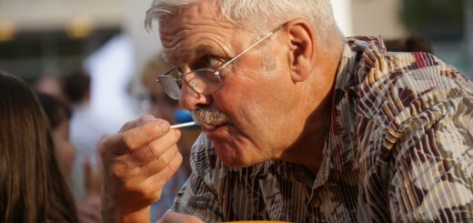 An older man sits at a table hugging a brown plate to his chest. He's leaning toward the table and sucking on a sucker stick as he looks over his glasses at someone. He has short gray hair in a classic cut ad wears a brown plaid shirt.