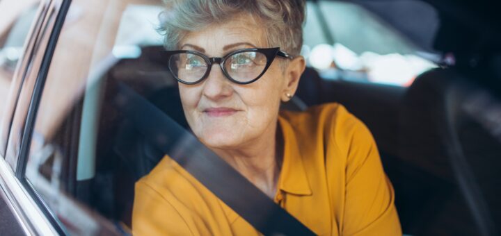 We see an older woman with short, spikey graying hair and thick-lensed glasses, looking out the side window of a car. She's wearing a gold collared top and has her seatbelt fastened.