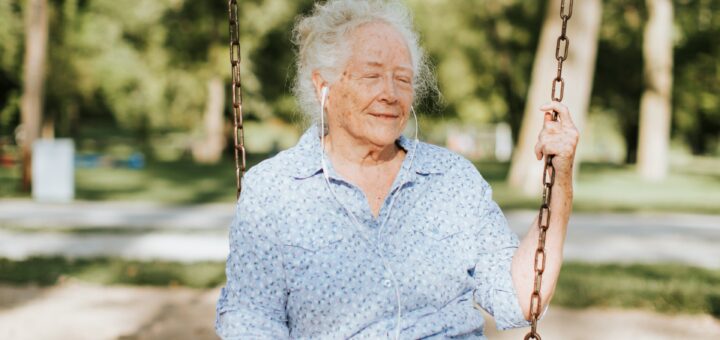 A woman sits happily in a bucket swing, eyes closed and listening to music through white earbuds. There are trees in the backgroud and she wears a light blue, open collared shirt and jeans.