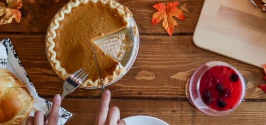 We see a dark wood, planked tabletop from the top view. Two hands are visible from a corner. On holds a fork, tines down, ready to spear a piece of pumpking pie. the pie has one slice missing. On the right, we see a dark veverage in a clear cup. Some decorative autumn leaves are scattered on teh table.