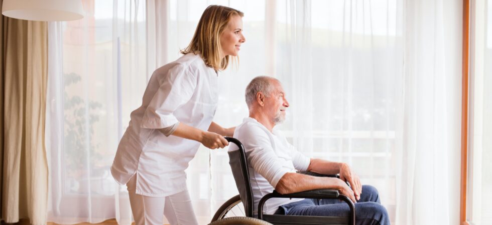 We see an older man wearing a white tee and drk pants sitting in a whieel chair. A pleasant looking young woman has her hands on the chair handles as if steaing him. She learns forard slightly as if speaking to him. Her clothing looks like white scrubs. Behind them are white sheers drawn over large windows.