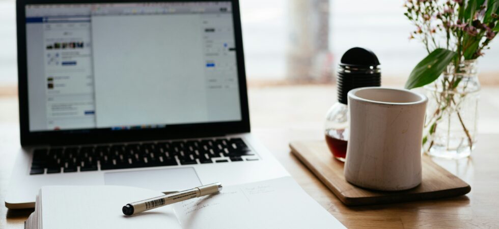 We view a laptop sitting open on a desk with a blank journal open in front. A pen lays on top and a cup of coffee or tea sits to the side. In the background, we see through a window to blurry greenry.