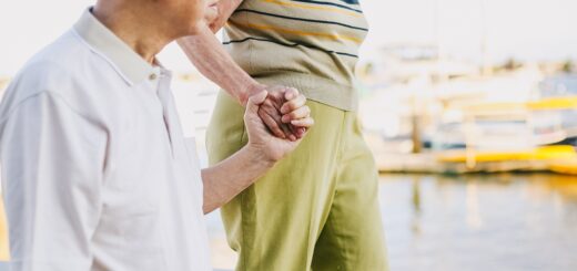 We see water behind as an older couple walks together holding hands. The woman appears to be on a higher elevation the the man. She wears a striped hsort sleepve top with lime green pants. She also has a white sun hat on. The man has gray hair and is wearing sun glasses perched on his head. He wears a white, short-sleeved button down and dark pants. The woman is looking ahead and the man is looiking out on the water at buildings across a bay area.