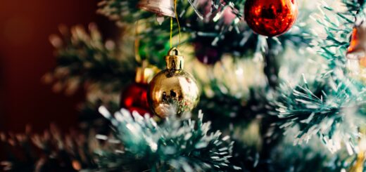 We see a very close up view of aportion of a traditionally decorated Christmas tree. A red bulb hangs from a branch in the top right corner ad scattered about se see a gold bell, gold bulbs and other ornaments.