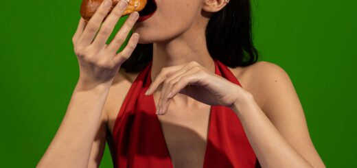 A young woman with darkhair combed back wears pink heart-shaped sunglasses and a red halter top. She's holding an ice cream confection to her mouth ready to take a bit. On a plate in front of her are more treats.