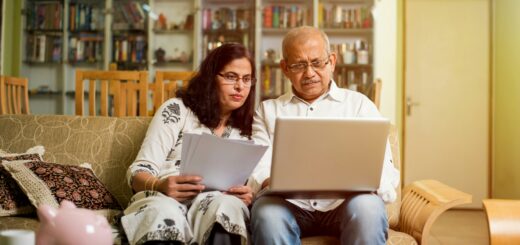 Father and daughter sit together looking at images or text on their laptops literally held on their laps. The older man wears jeans and a white long sleeved shirt. The younger woman wears a white top and pants with a black pattern.