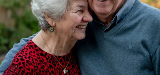 An older couple stand together, with the man holding the woman against him. They are both smiling as he draws her to his side. They both have gray hair. The oman wears a cranberry sweater tne the man wears a gray shirt.