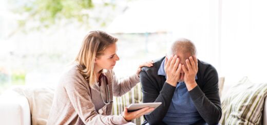 A younger woman sits to besides an older amn as whe trieds to comoft him. She has one hand on his shoulder as she learns forward talking to him. She wears a tan cardigan and white slacks. The man has his face buried in his hands, but we see the top of his head which has gray hair. He's wearing a navy blue cardigan over a lighter blue shirt.