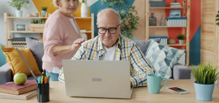 An older man wearing a plaind shirt and glasses sits at an open laptop, coffee cup at hand. Behind him, an older woman wearing a pink sweater, turns her had to talk with him.