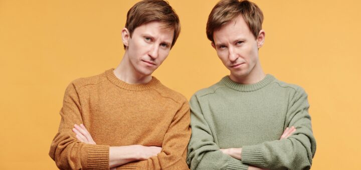 Two similar looking men, maybe twins, stand with their arms folded in front of them and angry looks on their faces. One wears a rust colored long-sleeved shirt, and the other wear the same shirt in sage green. The overall feeling is on of stubbornness.