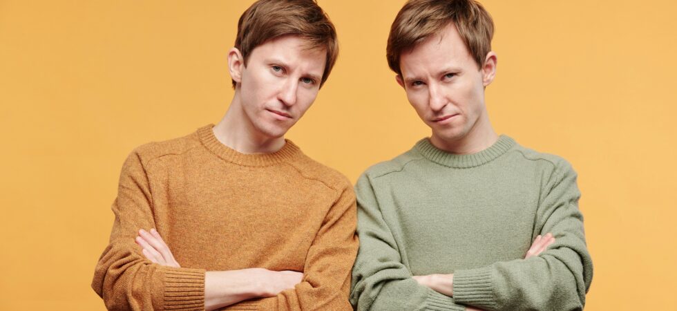 Two similar looking men, maybe twins, stand with their arms folded in front of them and angry looks on their faces. One wears a rust colored long-sleeved shirt, and the other wear the same shirt in sage green. The overall feeling is on of stubbornness.