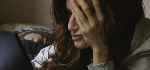 A tired lookin woman with plentiful dark hair holds a hand against her temple while she stares at a computer screen. The room is dark, as if evening, and she looks unable to do more.