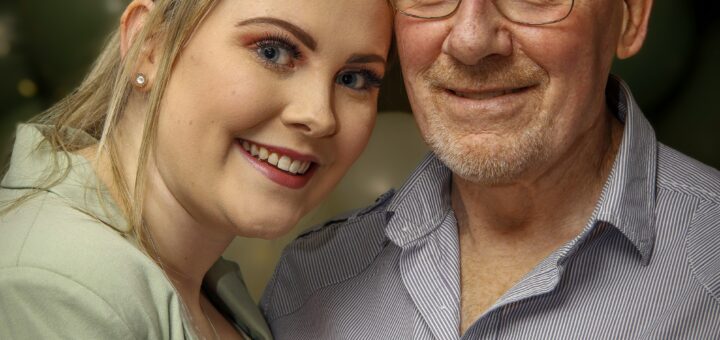 An young woman stands close to an older man, leaning slighly against him. The man is bald and wears glasses and a gray shirt. The young woman has medium brown hair pulled back with strands in front and wears a green sweater. They are both smiling.