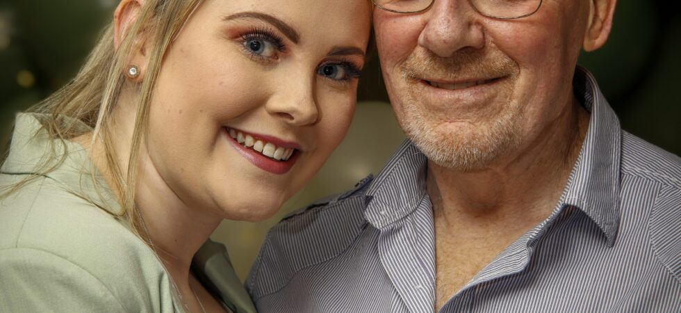 An young woman stands close to an older man, leaning slighly against him. The man is bald and wears glasses and a gray shirt. The young woman has medium brown hair pulled back with strands in front and wears a green sweater. They are both smiling.