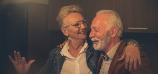 An older man with white hair and a trim beard and mustache sits on a couch next to an older woman. They appear to be sharing a laugh. The woman has gray hair pinned up, and wears glasses and a blue sweater. The man is wearing a brown cardigan.
