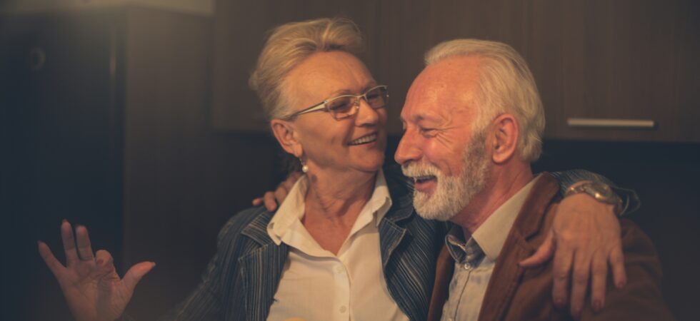 An older man with white hair and a trim beard and mustache sits on a couch next to an older woman. They appear to be sharing a laugh. The woman has gray hair pinned up, and wears glasses and a blue sweater. The man is wearing a brown cardigan.