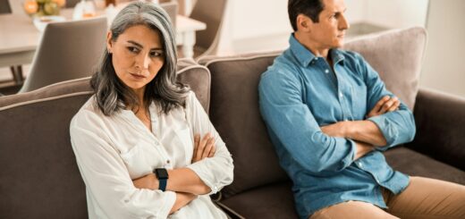 We see two middle-aged adults sitting at a distance on a couch, clearing aging. The woman on the left had her head turned away. She has shoulder length brown hair and is wearing a white shirt. Her arms are folded in defiance. The male on the right also has his head turned away and sits with arms folded. He has brown hair and is wearing a blue, long-sleeved shirt.