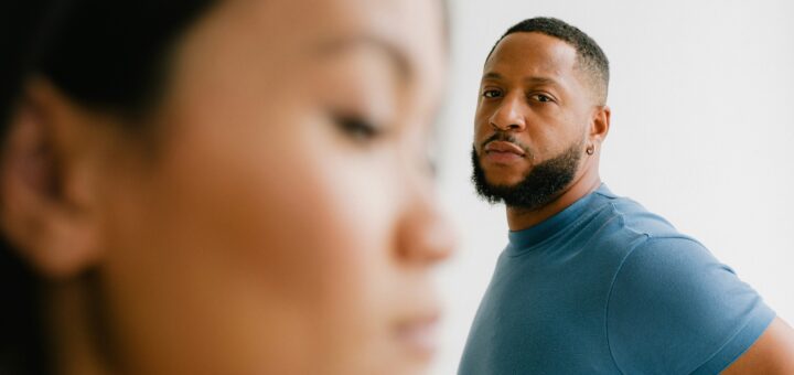 A slightly blurred woman's profile is in the forefront. She has coffee toned skin, and dark hair. We see her looking down as if downcast. In the background, we see a man with darker skin and a black chin beard with his head turned toward her with an angry expression. He wears a blue sweater.