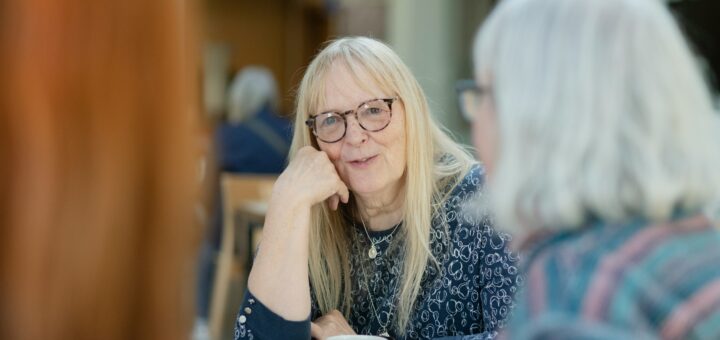 Two older women sit at a table set with white tea cups. The one facing the camera has long blonde/gray hair and glasses and sits with her elbow on the table, resting her chin on her hand. She's speaking to a woman we see in profile who has medium length white hair and glasses.