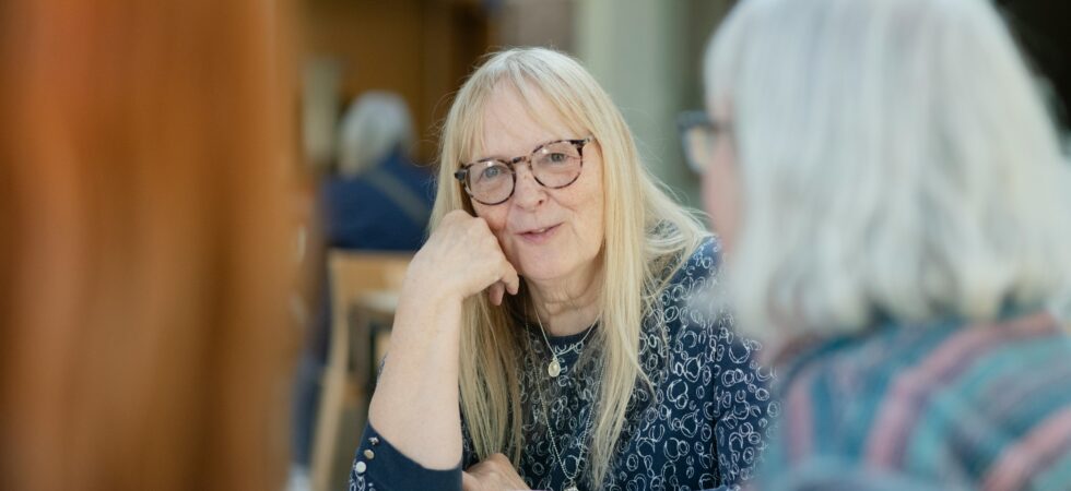 Two older women sit at a table set with white tea cups. The one facing the camera has long blonde/gray hair and glasses and sits with her elbow on the table, resting her chin on her hand. She's speaking to a woman we see in profile who has medium length white hair and glasses.