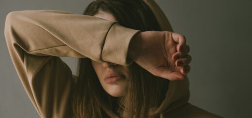 A woman with long dark hair holds her arm up over her eyes as if wanting to avoid what's in front of her. She's wearing a tan hoodie.