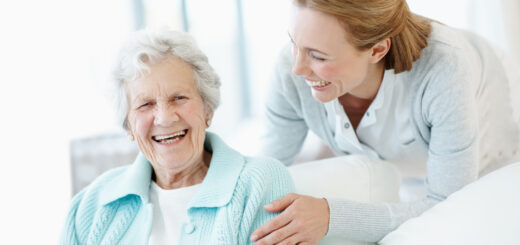 And older woman with white hair sits in a chair with a younger woman in a medical coat leaning over her, touching her shoulder. They are both smiling.