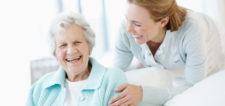 And older woman with white hair sits in a chair with a younger woman in a medical coat leaning over her, touching her shoulder. They are both smiling.