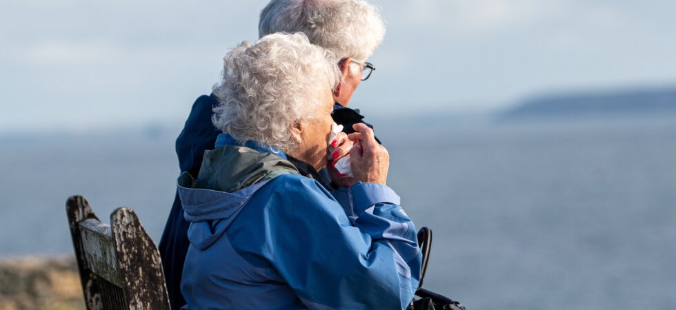 An older couple sites side by side on a bench, looking out over a lake.
