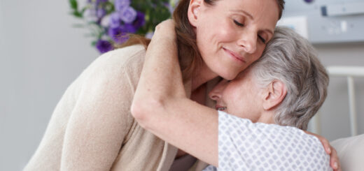 An older woman with gray hair is hugging a younger woman who is bending over her. The older woman is wearing a hospital gown and the younger woman has long dark hair and is wearing a beige sweater.