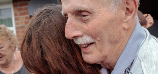 An older man with gray hair and a gray mustache is seen in profile as he hugs a younger woman seen from the back.