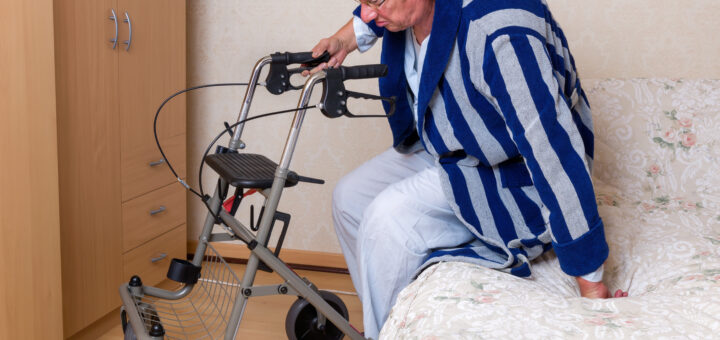 An older man in a striped bathrobe attempts to balance himself on his rollator as he struggles to get up off the bed.
