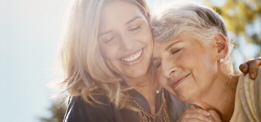 We see a younger woman with blond hair embrace an older, white-haired woman from the side. The older woman lays her head against the younger woman's chest. They are both smiling.