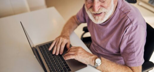 This image shows a laptop on a white surface. An older man with neat white hair and a trim beard looks up at the camera. He's wearing a pink short-sleeved knit shirt.
