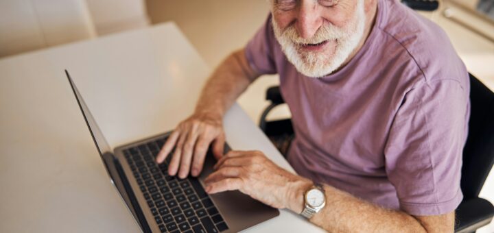 This image shows a laptop on a white surface. An older man with neat white hair and a trim beard looks up at the camera. He's wearing a pink short-sleeved knit shirt.