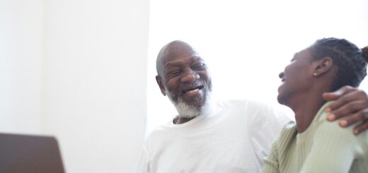 A older man sits at a computer at a desk. His head is turned as he smiles at a younger woman whom he obviously loves. His arm is reaching around her shoulders. Both have dark skin; the man has gray hair and a goatee.