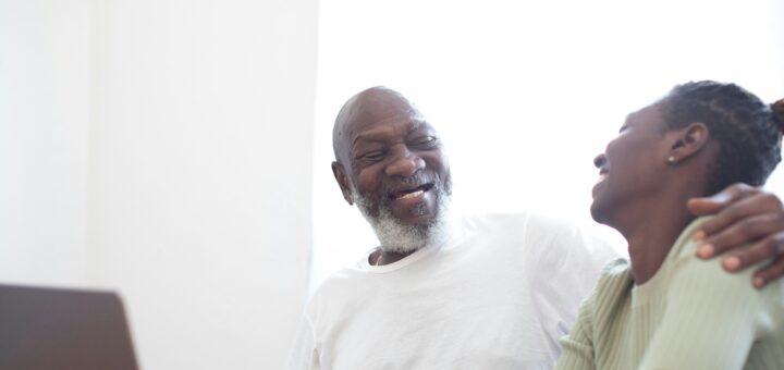 A older man sits at a computer at a desk. His head is turned as he smiles at a younger woman whom he obviously loves. His arm is reaching around her shoulders. Both have dark skin; the man has gray hair and a goatee.