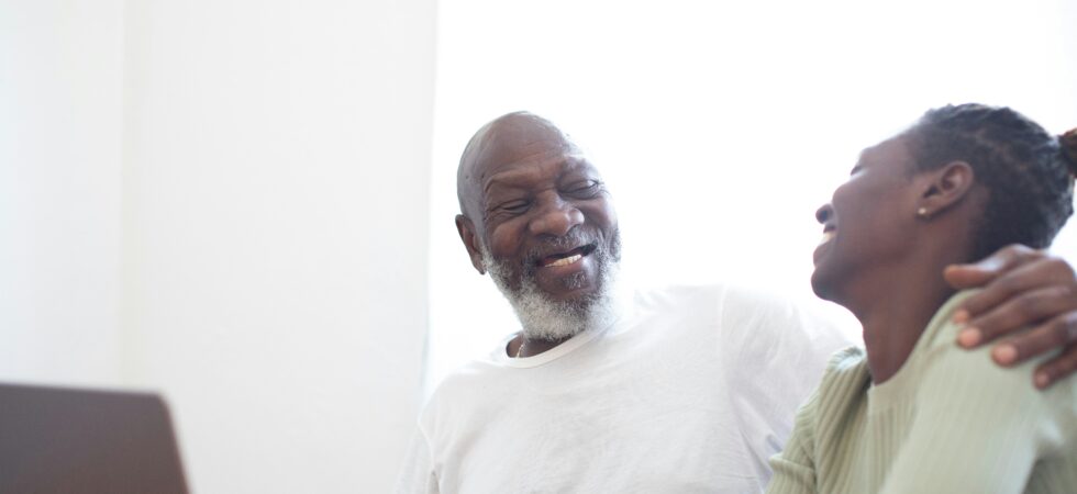 A older man sits at a computer at a desk. His head is turned as he smiles at a younger woman whom he obviously loves. His arm is reaching around her shoulders. Both have dark skin; the man has gray hair and a goatee.