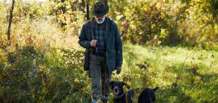 An older man wearing hunting gear walks with his dogs at his side. He's wearing a hunting cap, jacket and jeans. and has a shotgun over his shoulder.