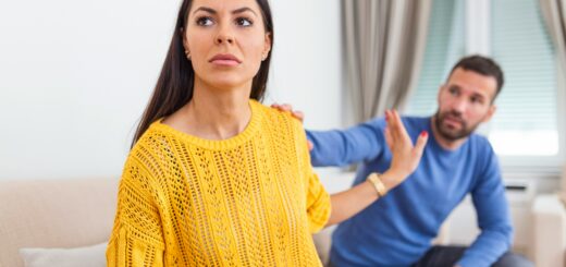 A woman in a bright gold sweater sits on a couch with her back turned to a man, hand help out to keep him at bay. The man looks at her as if arguing. Both are upset.