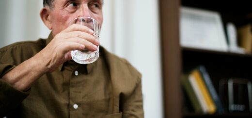 An older man looking slightly confused looks up at someone as he lifts a glass of water to his lips. He wears a brown button-up shirt.