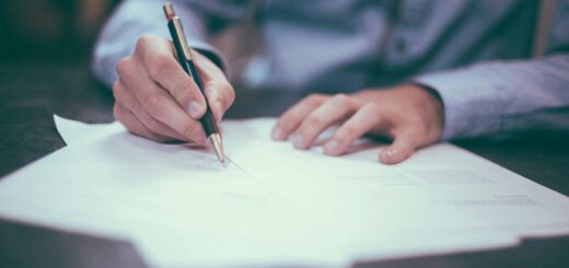 A man sits at a desk signing official looking papers. We only see his arms and torso clad in a light blue dress shirt as he holds a pen and signs.